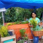 Na imagem, uma mulher de blusa verde segura uma cesta com plantas medicinais em frente a uma barraca laranja que tem outras plantas e bananas